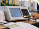 Urna eletrônica brasileira em local de votação, com eleitor confirmando o voto e elementos de auditoria ao redor